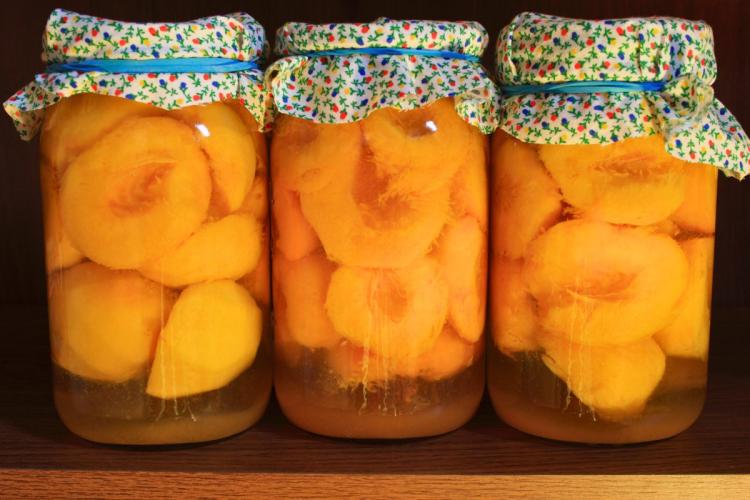 Three jars of sliced canned peaches, home canned, submerged in liquid, with decorative floral cloth covers on the lids.