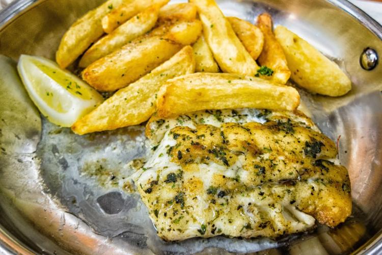 Grilled kingklip fish seasoned with herbs, served alongside thick-cut golden fries and a lemon wedge on a metal plate.