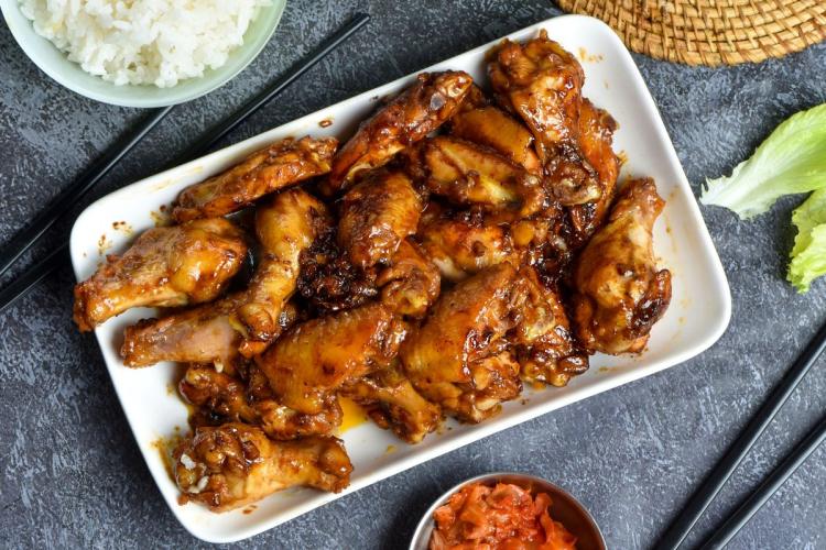 Plate of honey soy-glazed chicken wings with a glossy dark brown glaze, served alongside a bowl of white rice and a small portion of pickled vegetables, with chopsticks and lettuce as part of the setting.
