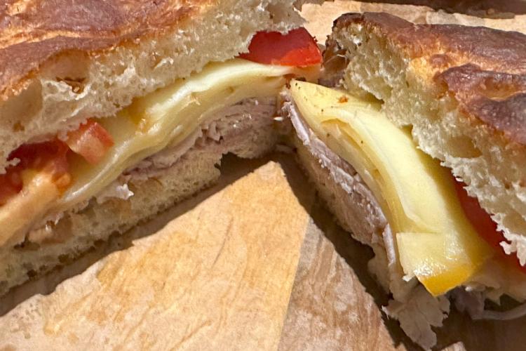 Close-up of a sandwich with rustic bread, porchetta, cheese, and sliced tomatoes.