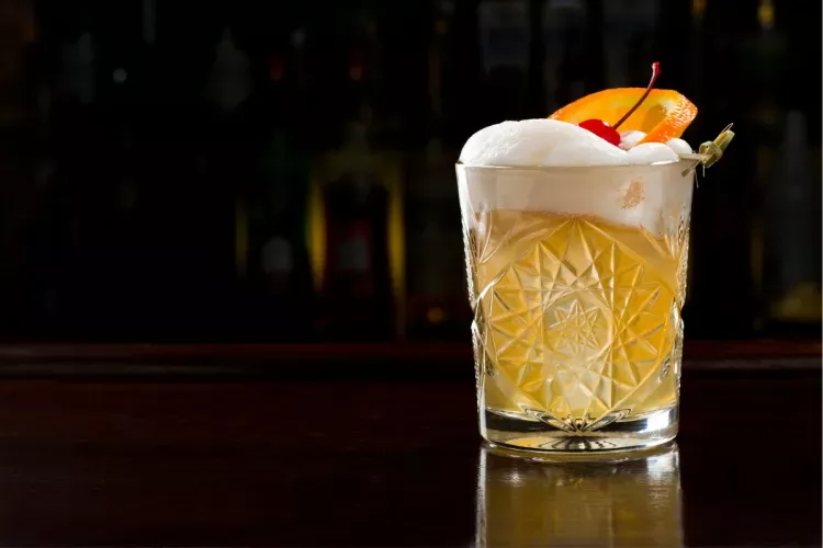 Classic whiskey sour in a patterned glass with a frothy egg white top, garnished with an orange slice and a cherry, set against a dimly lit bar background.