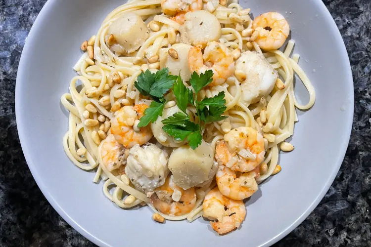 Linguine pasta Freeport, topped with shrimp, scallops, toasted pine nuts, and a sprig of parsley on a gray plate.