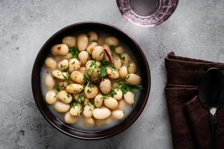 Bowl of white bean stew in broth, topped with fresh herbs, next to a brown napkin and spoon on a gray surface.