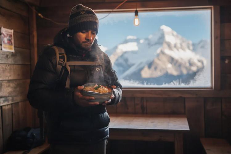 A trekker sits inside a cozy teahouse, holding a steaming bowl of thukpa, a popular Nepali dish made with noodles, vegetables, and spices, while snowy mountains are visible through the window, showcasing the breathtaking landscape of Nepal. The scene captures the essence of Nepali cuisine and the warmth of local hospitality amidst the majestic Himalayas.