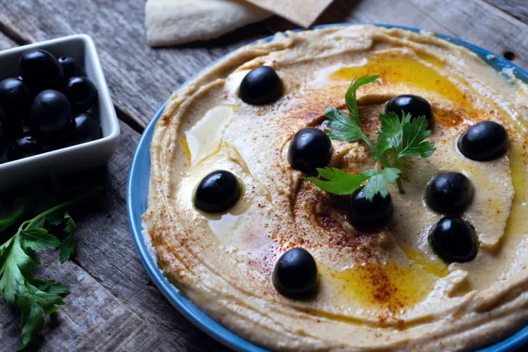 Plate of swirled hummus with olive oil, paprika, and black olives, with pita bread and a bowl of olives nearby.