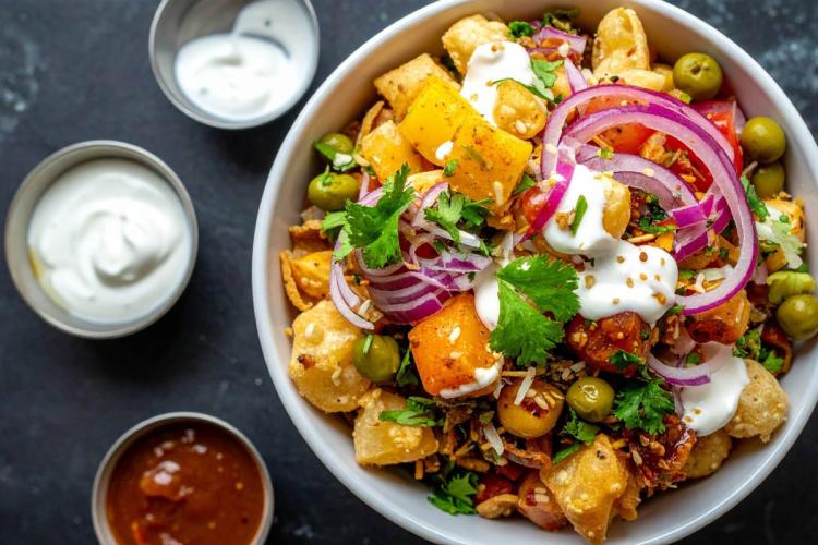 A bowl of aloo chaat with spiced fried potato cubes, green peas, red onion, tomato, papdi, cilantro, and a yogurt drizzle, served alongside small bowls of raita and tamarind chutney on a dark surface.
