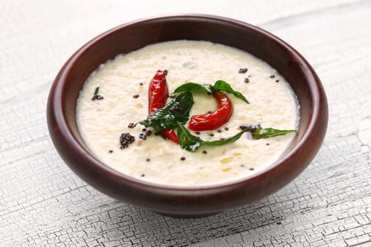 A clay bowl of smooth white coconut chutney garnished with a tempering of dried red chilies, curry leaves, and black mustard seeds, placed on a rustic white wooden surface.