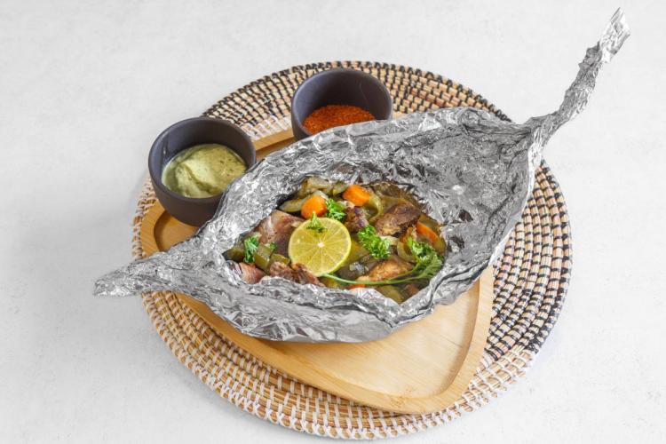 Foil-wrapped fish with vegetables and lime served on a wooden tray with dipping sauces, Congolese style