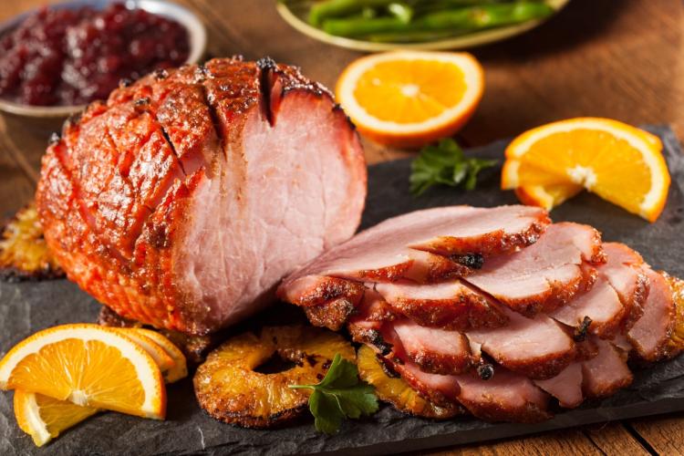 Sliced glazed baked ham served with orange wedges and grilled pineapple rings on a dark serving board.