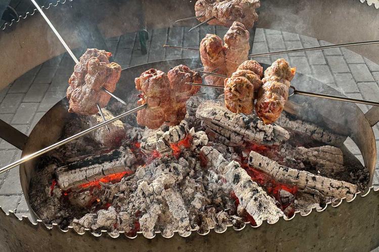 Several rolled meat roasts on metal spits cooking over glowing embers and burning logs in a large circular open-fire grill, with smoke rising from the coals.