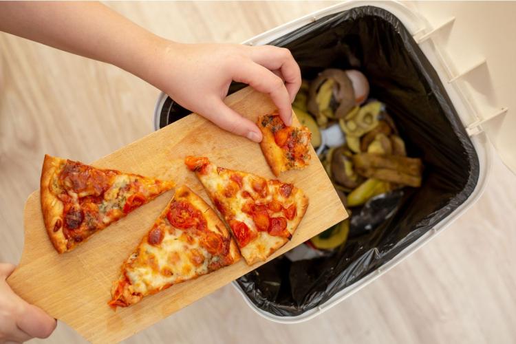 A hand scraping three uneaten slices of tomato pizza off a wooden board into a kitchen bin already filled with discarded vegetable peels and food scraps.