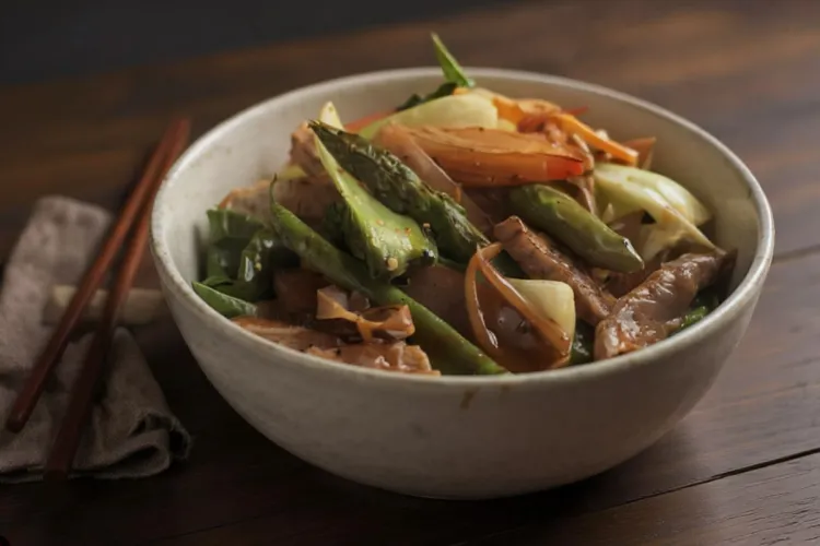 A pale stoneware bowl of pork and vegetable stir-fry — strips of pork with green asparagus, flat green beans, onion wedges, and red bell pepper in a light glossy sauce — on a dark wood table with chopsticks resting on a linen napkin. Synthetic (AI-generated) image.