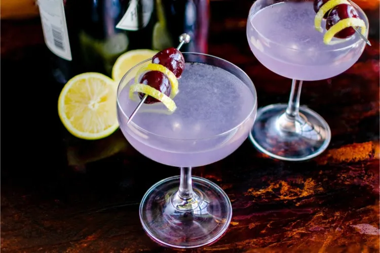 Two pale lavender cocktails in coupe glasses, each garnished with a cherry and a lemon twist on a pick, with a halved lemon and bottles behind on a dark surface.