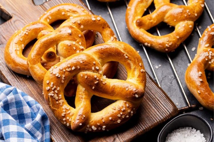Freshly baked Bavarian pretzels scattered with coarse salt on a dark wooden board, with more cooling on a wire rack and a bowl of salt alongside