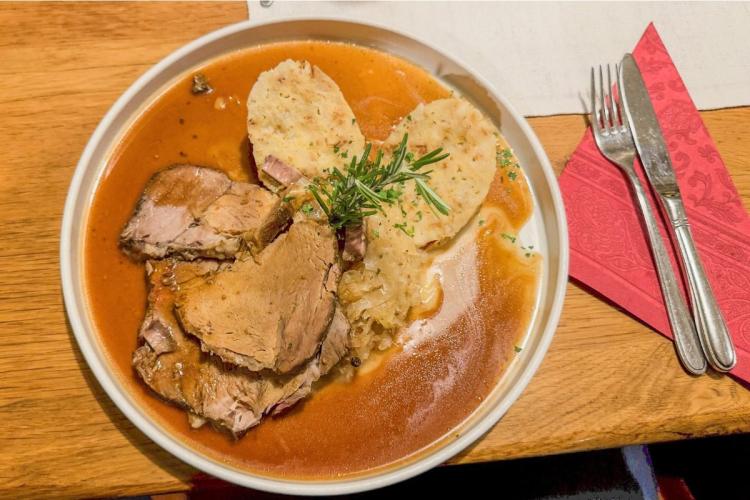 Bowl of sliced Bavarian roast pork in dark gravy with two bread dumplings, sauerkraut and a rosemary sprig, on a wooden table beside a red napkin and cutlery