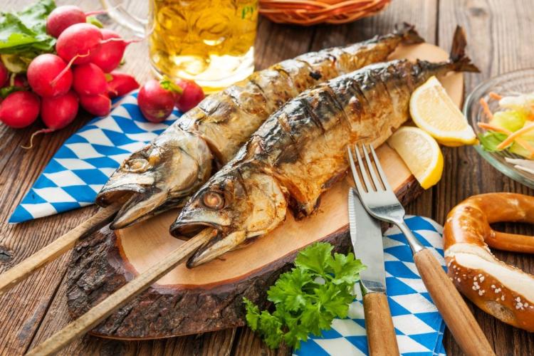 Two whole grilled mackerel on wooden skewers resting on a bark-edged board with lemon wedges, radishes, parsley, a pretzel and a glass of beer