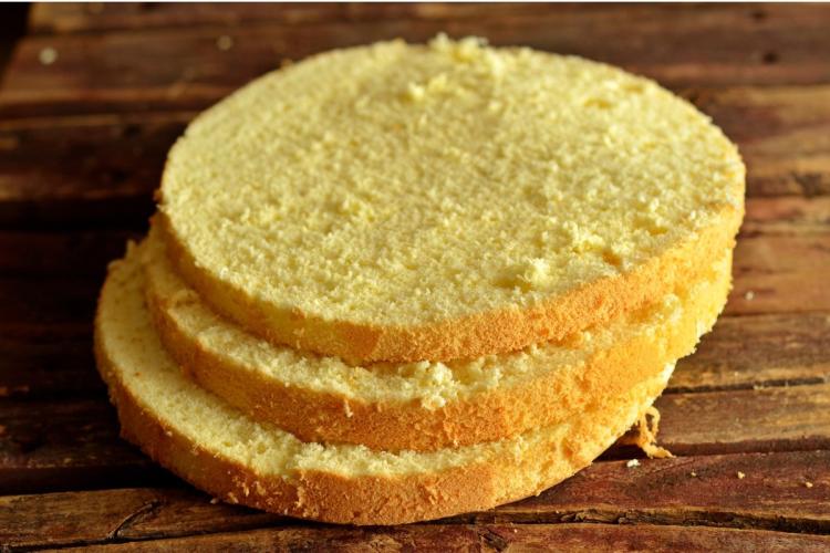 Three slices of a pale, fine-crumbed gluten-free sponge cake stacked on a rustic wooden board