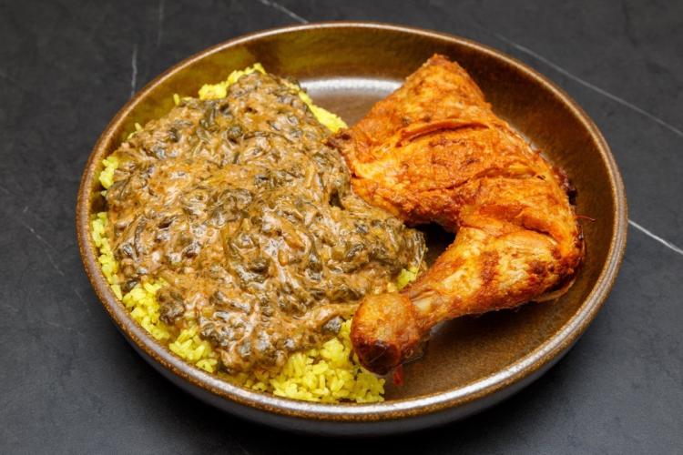 A plate of spinach mwamba, a creamy peanut and spinach sauce, spooned over yellow rice and served with a roasted chicken leg, on a dark stoneware plate