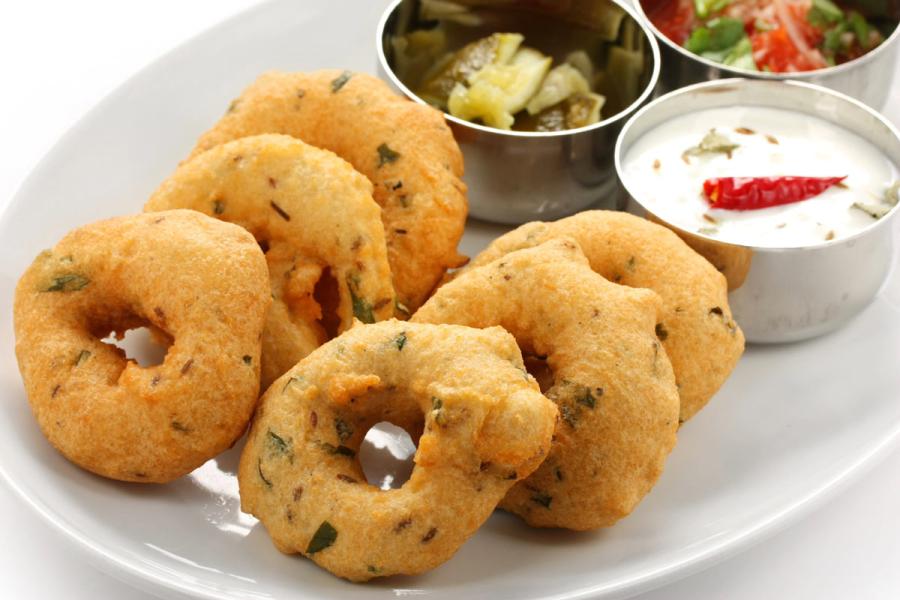 Six golden medu vada, South Indian lentil fritters, on a white plate, served with three small bowls of green chutney, tomato salsa, and raita.