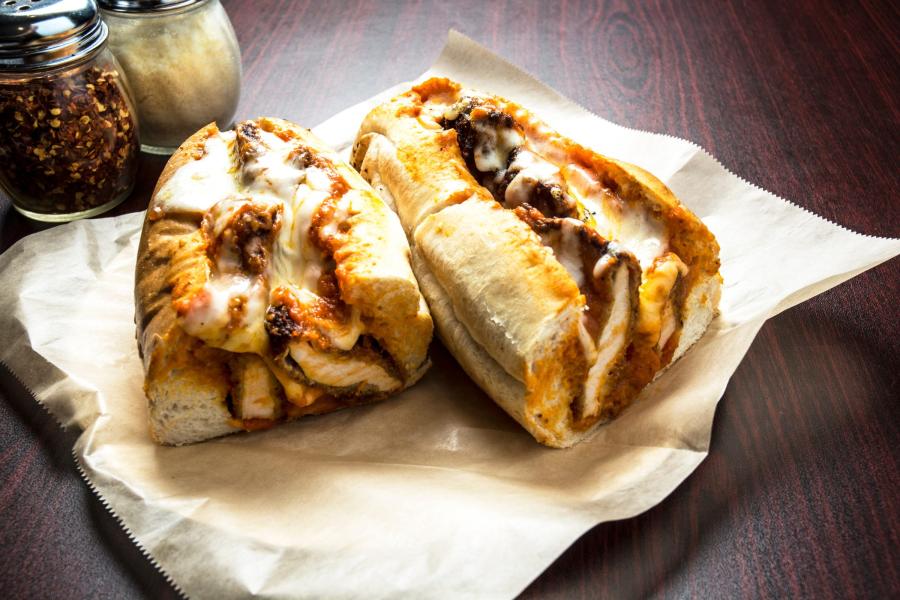 Chicken Parmesan hero sandwich cut in half, with breaded chicken cutlets, melted mozzarella cheese, and marinara sauce in a crusty hoagie roll, placed on parchment paper. Jars of grated Parmesan cheese and crushed red pepper flakes are in the background on a dark wooden table.