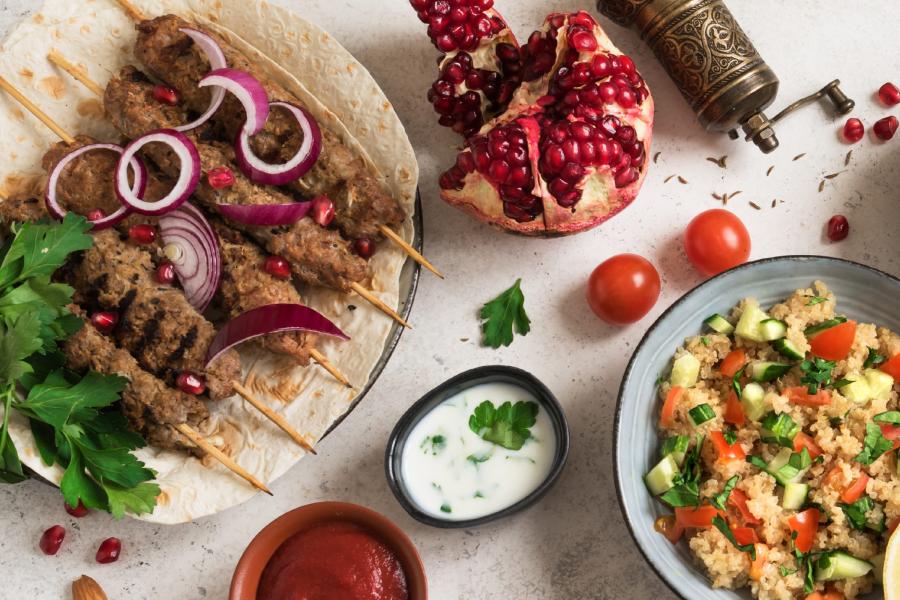 Middle Eastern meal with grilled meat kebabs on skewers garnished with red onion and pomegranate seeds, served on flatbread. Accompanied by a bowl of couscous salad with cucumber, tomato, and herbs. Additional garnishes include parsley, a halved pomegranate, cherry tomatoes, and small bowls of yogurt sauce and red sauce.