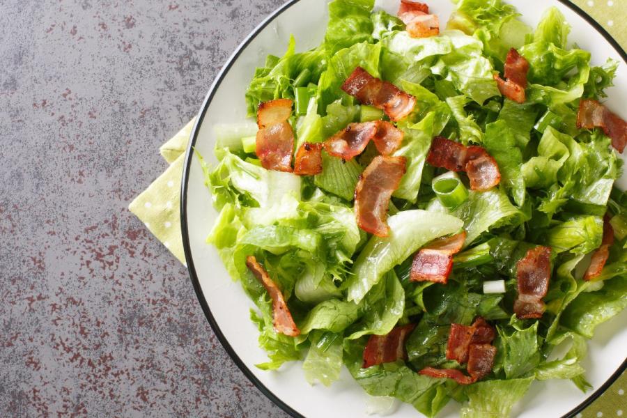 Salad with lettuce and crispy bacon pieces in a white dish with a black rim, placed on a stone-like countertop with a light green polka-dotted napkin underneath.
