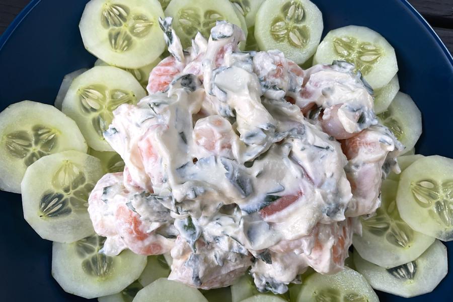 A prawn salad with creamy dressing and herbs, surrounded by neatly arranged cucumber slices on a dark blue plate.