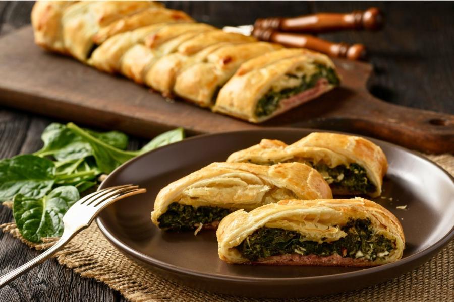 Close-up of a puff pastry pie filled with spinach, ham, and cheese, sliced on a dark brown plate with fresh spinach leaves on the table and the remaining pie on a wooden cutting board in the background.