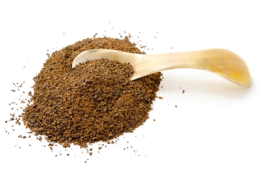 A pile of dark brown ground wattleseed with a small scoop on a white surface.