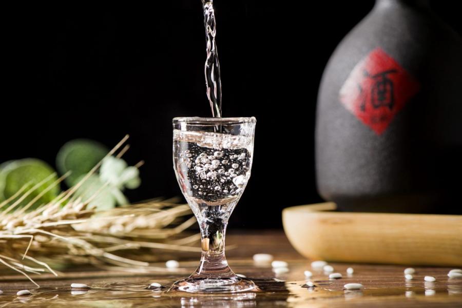 A clear bajiu being poured into a small glass with bubbles forming, with a ceramic bottle and scattered rice grains in the background.