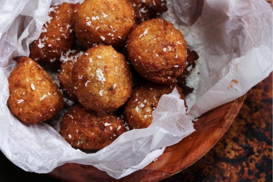 Wooden bowl filled with golden-brown, round banana fritters, garnished with shredded coconut and lined with parchment paper.
