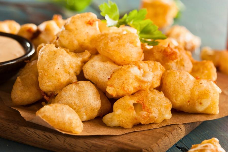 Golden, crispy battered cheese curds served on a wooden board with parchment paper, accompanied by a creamy dipping sauce and garnished with fresh parsley.
