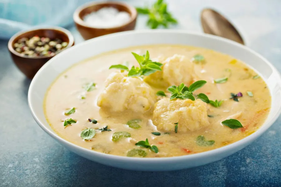 A bowl of creamy chicken soup with fluffy dumplings, garnished with fresh herbs, served on a blue surface with seasoning bowls and a wooden spoon in the background.