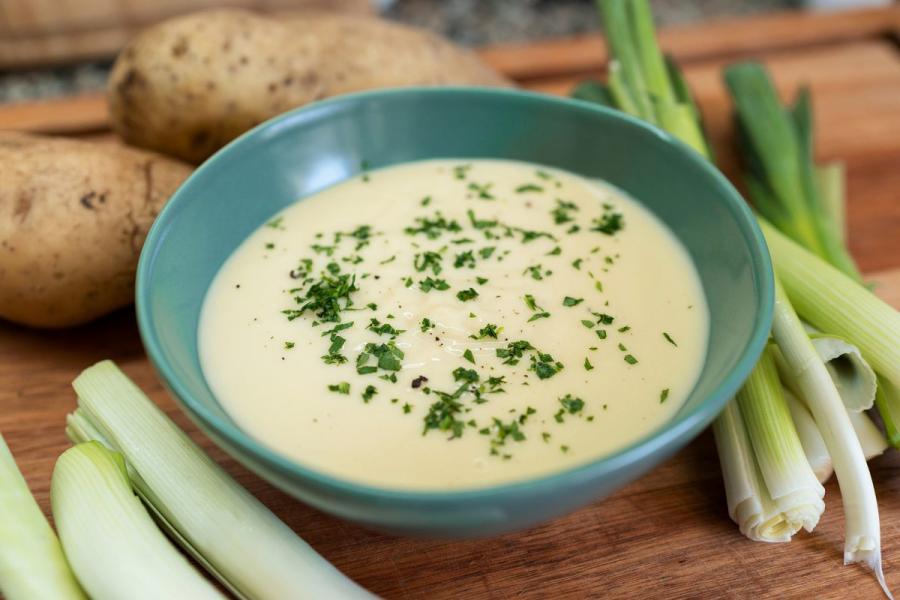 Close-up of a bowl of creamy vichyssoise soup garnished with fresh herbs, placed on a wooden board with raw leeks and potatoes in the background.