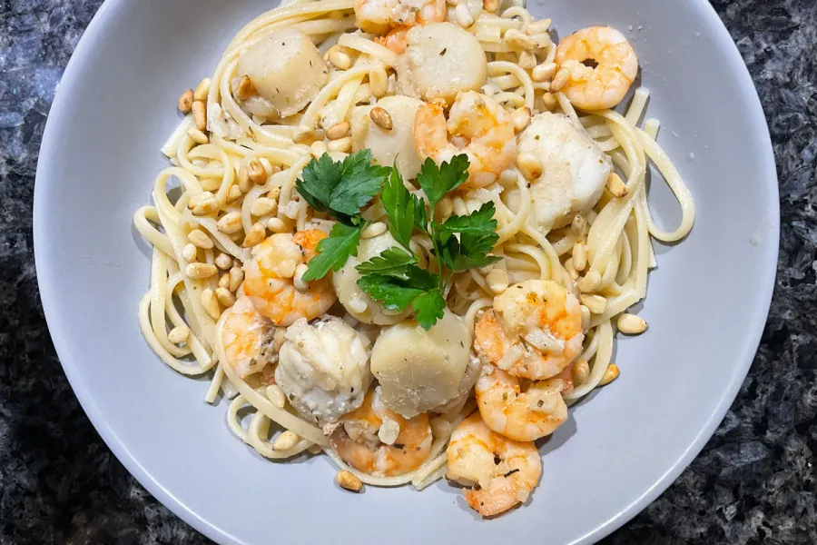 Linguine pasta Freeport, topped with shrimp, scallops, toasted pine nuts, and a sprig of parsley on a gray plate.
