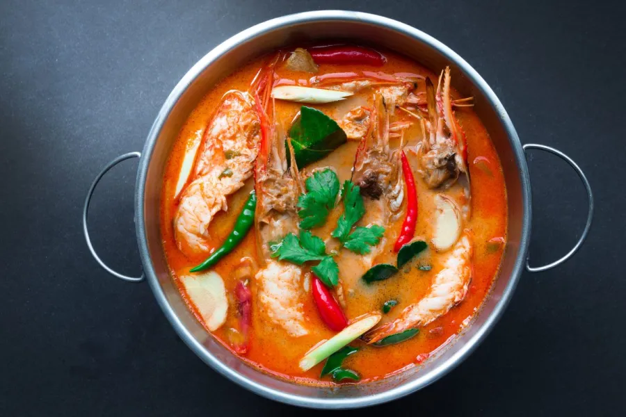 A pot of tom yum goong, Thai spicy and sour shrimp soup with prawns, herbs, and chilies in a bright orange broth.