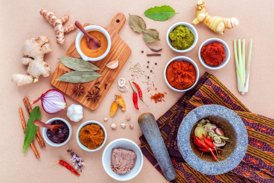 Flat lay of Thai cooking ingredients including fresh herbs, curry pastes, spices, and a stone mortar and pestle on a neutral background.