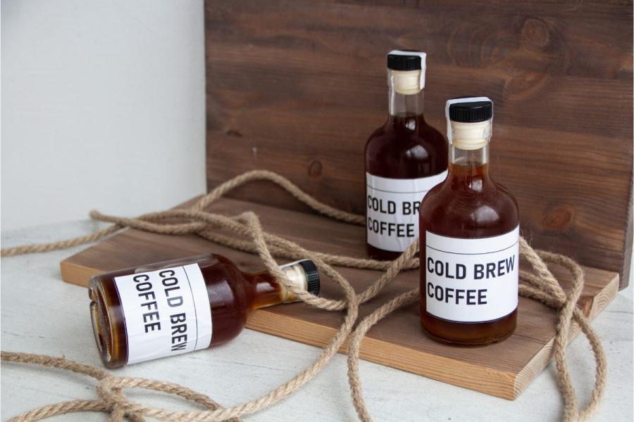 Three glass bottles of cold brew coffee with white labels reading 'COLD BREW COFFEE' arranged on a wooden board and light surface, with a natural fiber rope draped around them.