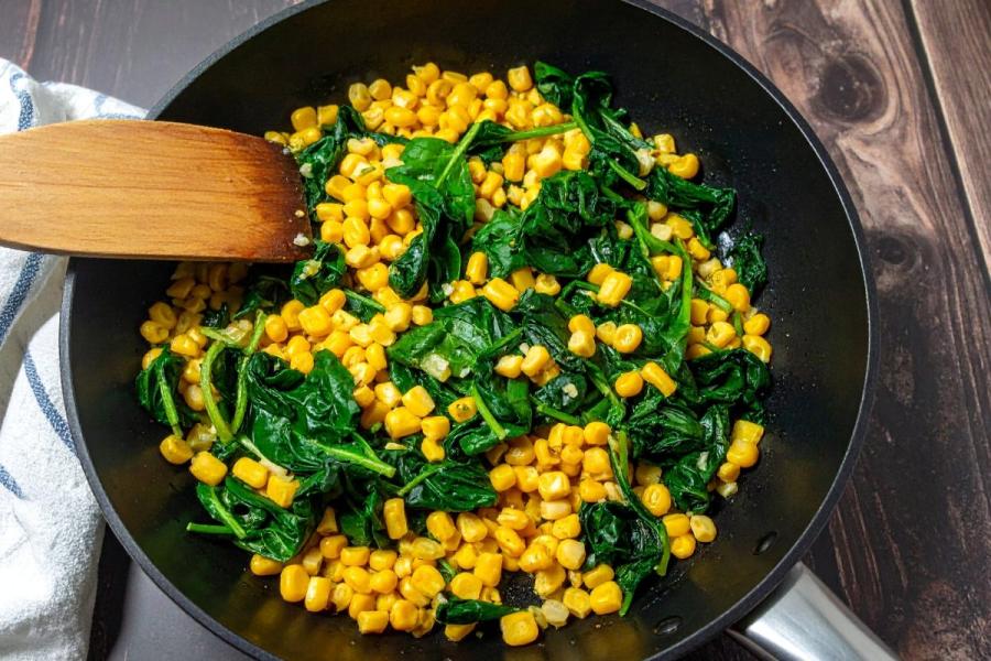 Skillet of sautéed corn and spinach with a wooden spatula on a dark wooden table.