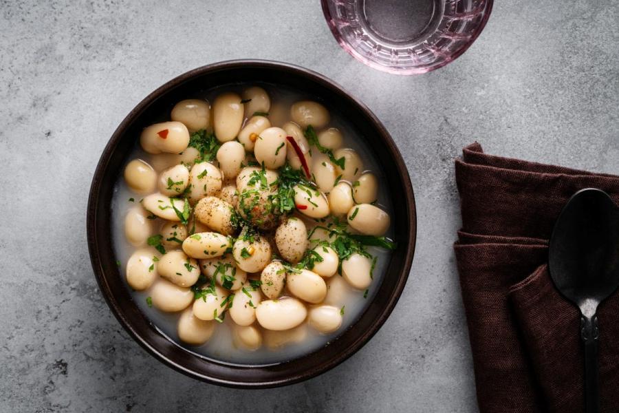 Bowl of white bean stew in broth, topped with fresh herbs, next to a brown napkin and spoon on a gray surface.