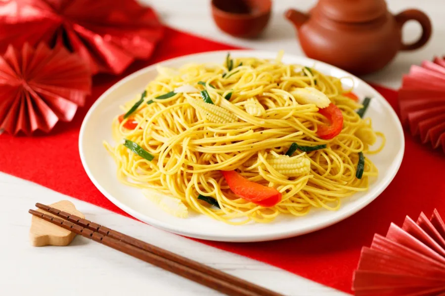 Longevity noodles stir-fried with baby corn, red bell pepper, and green onions on a white plate, with chopsticks and red Lunar New Year decorations.