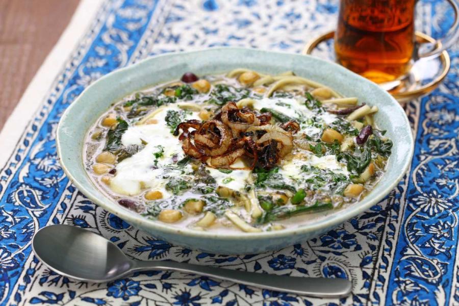 A plate of ash reshteh, a thick Persian herb, bean, and noodle soup, topped with white kashk and fried onions.
