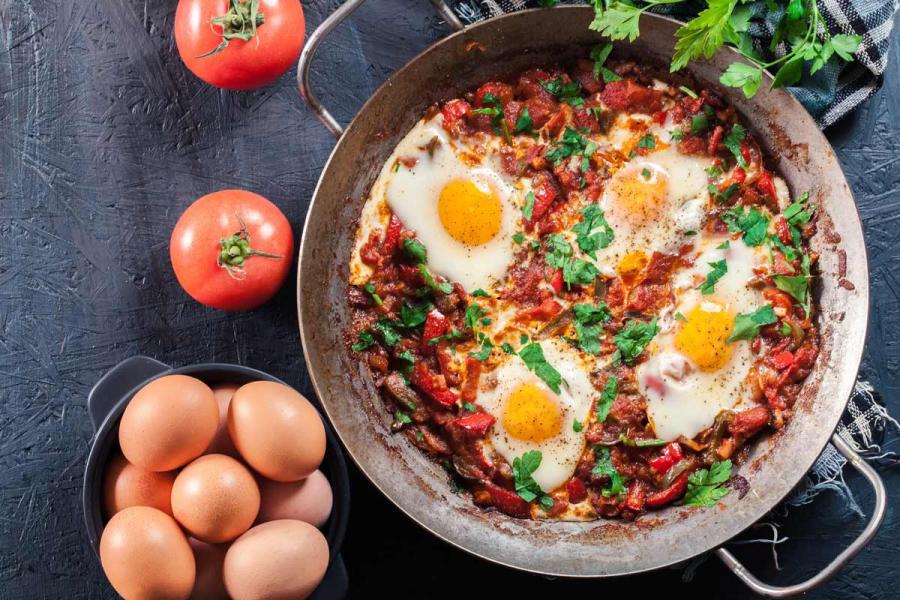 Classic shakshuka with four poached eggs in a spiced tomato and pepper sauce, garnished with fresh parsley, served in a steel pan on a dark slate surface with tomatoes and raw eggs alongside.