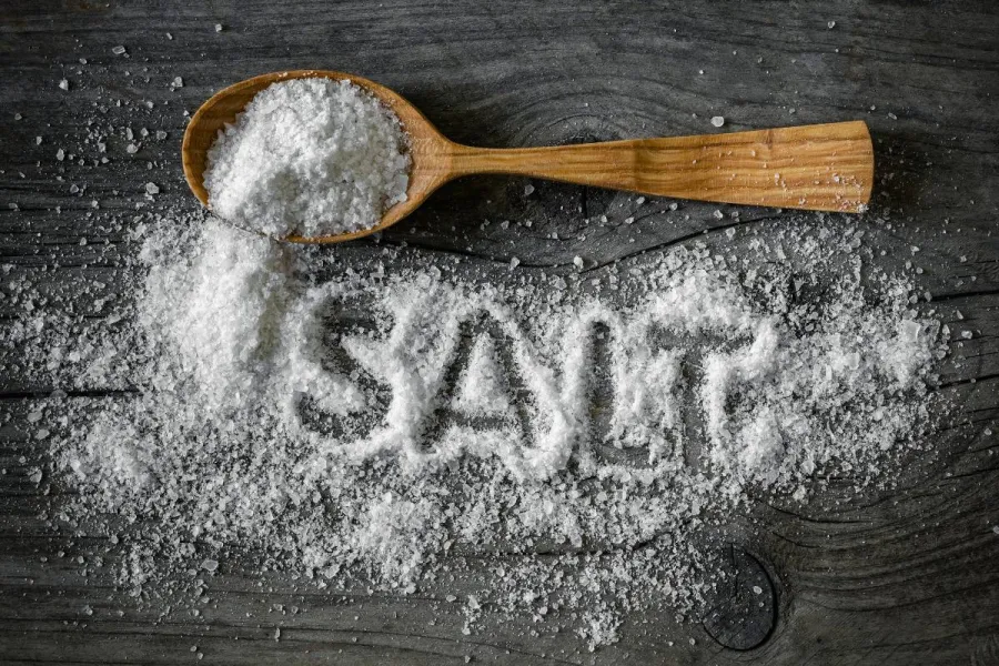 Wooden spoon filled with coarse salt on a dark table, with scattered salt spelling the word SALT.