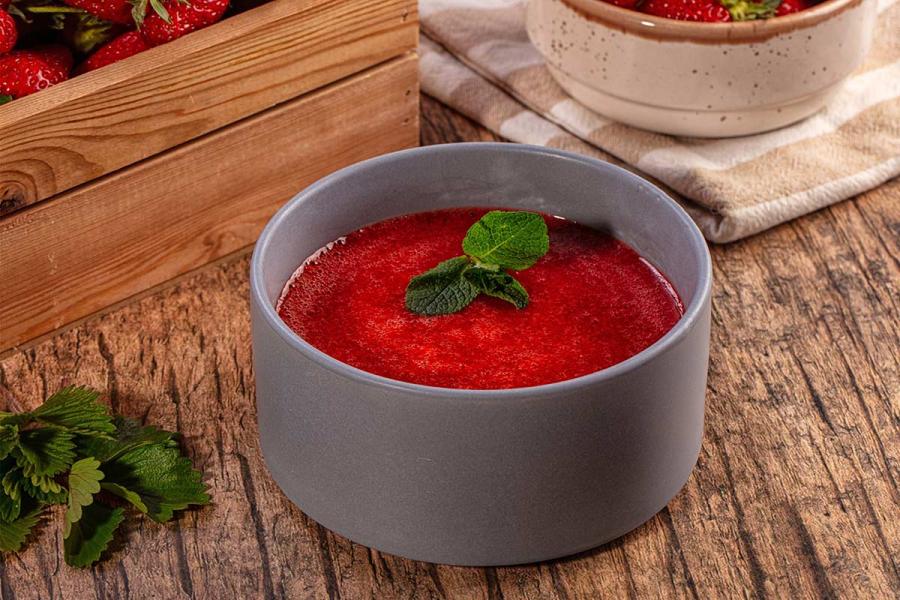 Gray bowl filled with smooth strawberry coulis topped with mint leaves on a rustic wooden table, with fresh strawberries in the background.