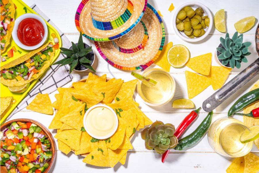 Overhead view of a festive Mexican-themed table with tortilla chips, tacos, salsa, lime drinks, peppers, sombreros, and colorful party decor.