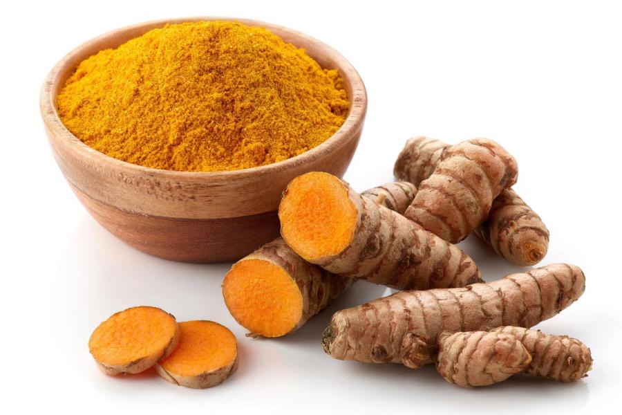 Wooden bowl of turmeric powder next to fresh turmeric roots and sliced turmeric showing bright orange flesh on a light background.