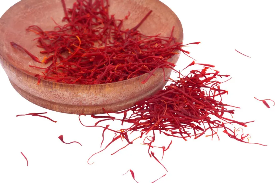 Red saffron threads in a wooden bowl with some spilled on a white background.