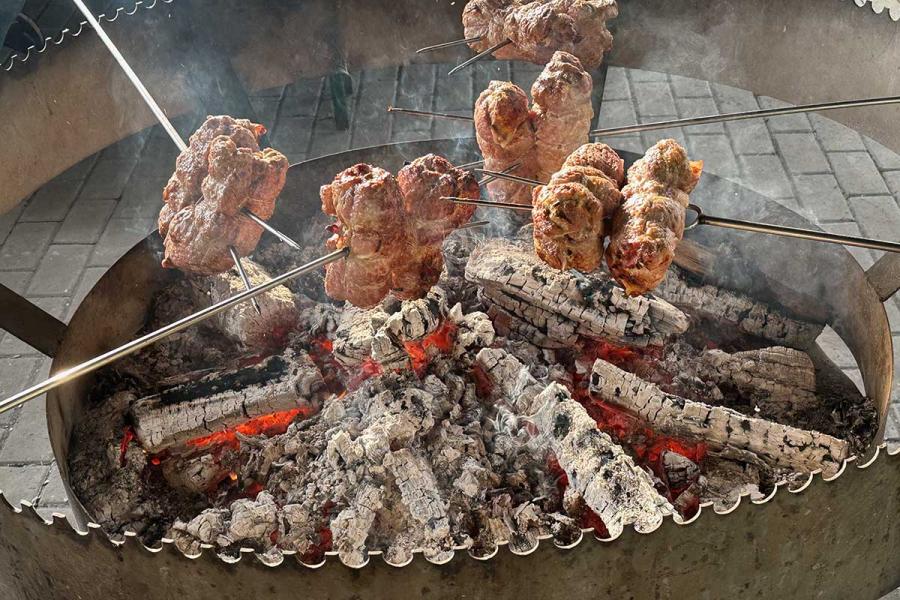 Several rolled meat roasts on metal spits cooking over glowing embers and burning logs in a large circular open-fire grill, with smoke rising from the coals.