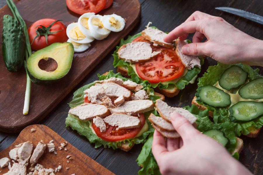 Person preparing turkey sandwiches with lettuce, tomato, cucumber, and sliced turkey on a dark wooden table, surrounded by fresh vegetables, avocado, and hard-boiled eggs.